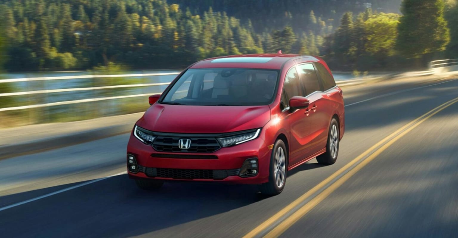 A red minivan driving on a road with a blurred background of trees and water.