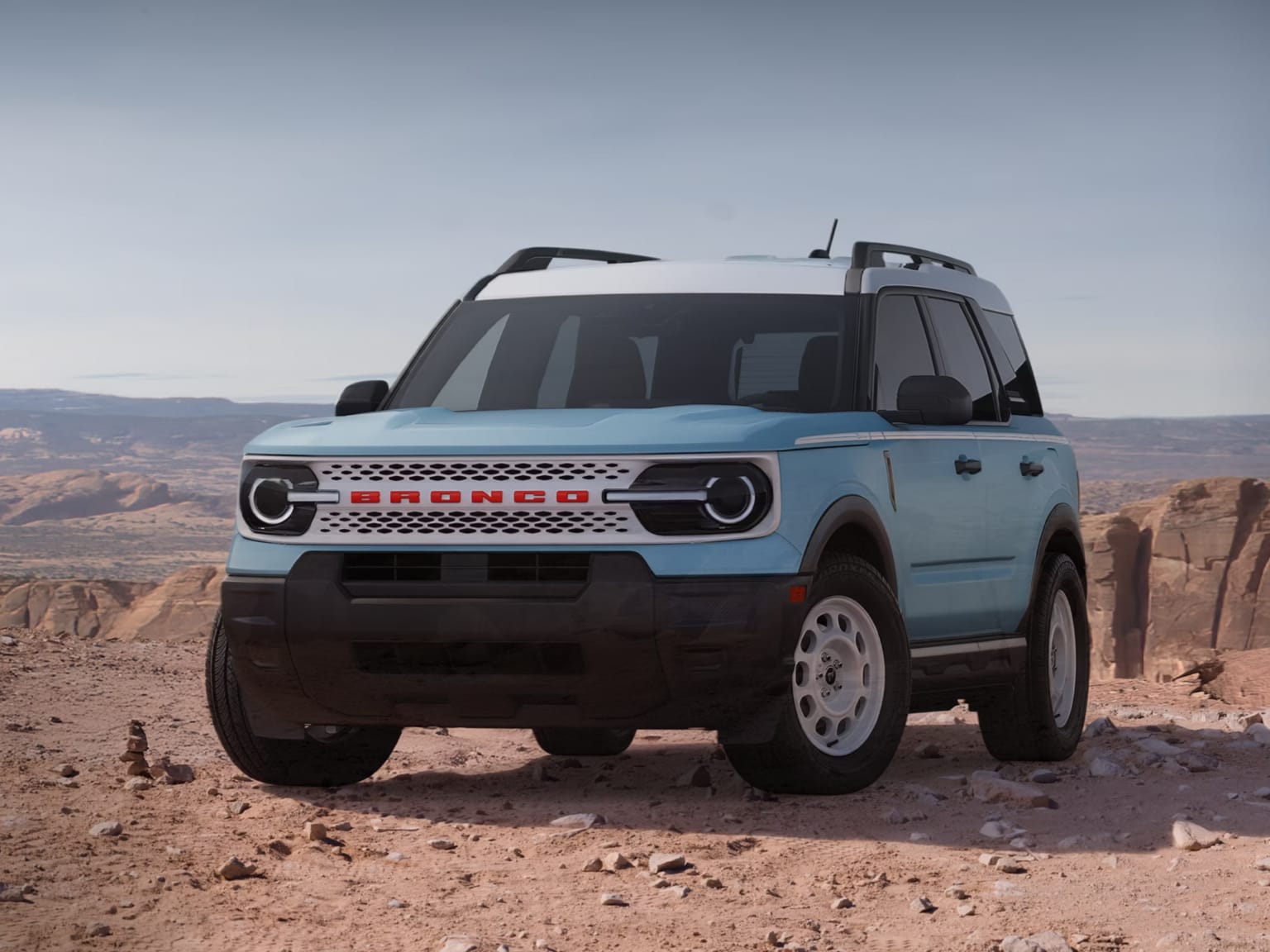 A light blue compact SUV sits on a rocky desert plateau against a backdrop of red sandstone formations and a clear sky.