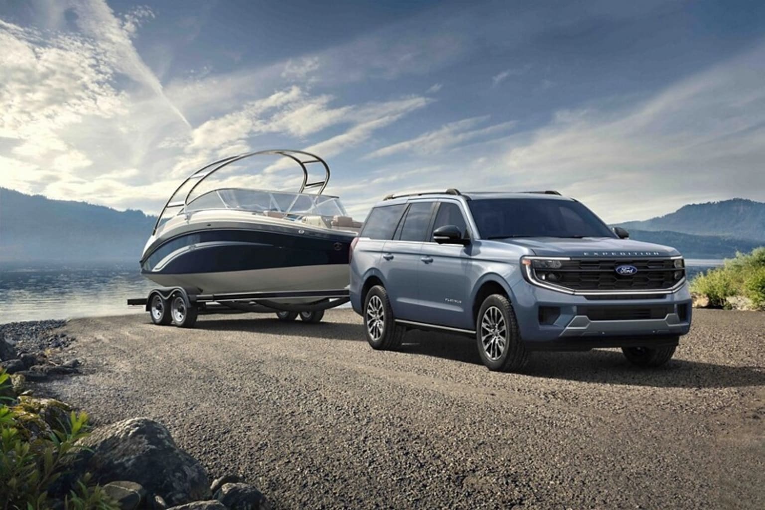 A large silver SUV towing a boat on a trailer, parked on a gravel path near a body of water surrounded by mountains and a cloudy sky.