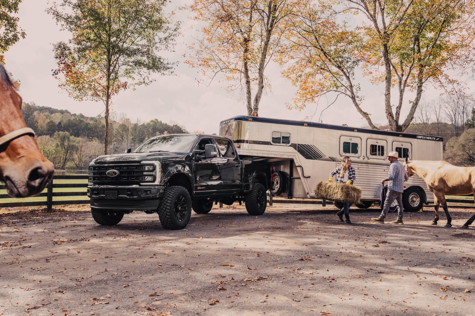 A large black pickup truck with oversized tires parked in a rural setting, surrounded by autumn-colored trees, with a horse-drawn trailer in the background.