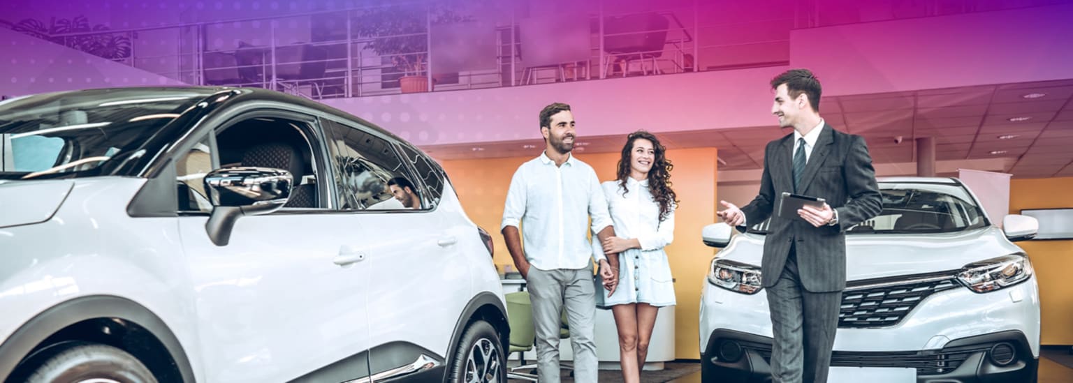 A couple walking between two white cars in a showroom while a salesman in a suit stands to the right, holding a tablet.