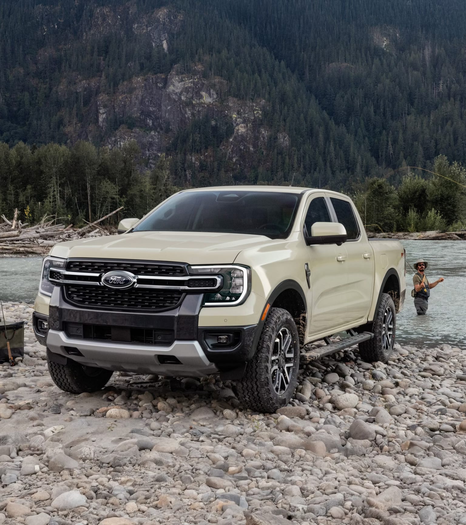 A tan pickup truck parked on a rocky riverbank with a fisherman in the water and forested mountains in the background.