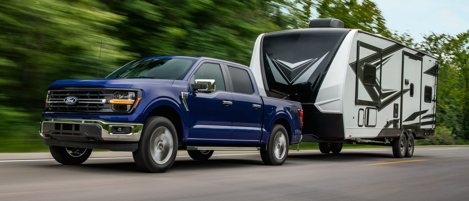 A blue pickup truck tows a white and black travel trailer down a road with blurred green trees in the background.