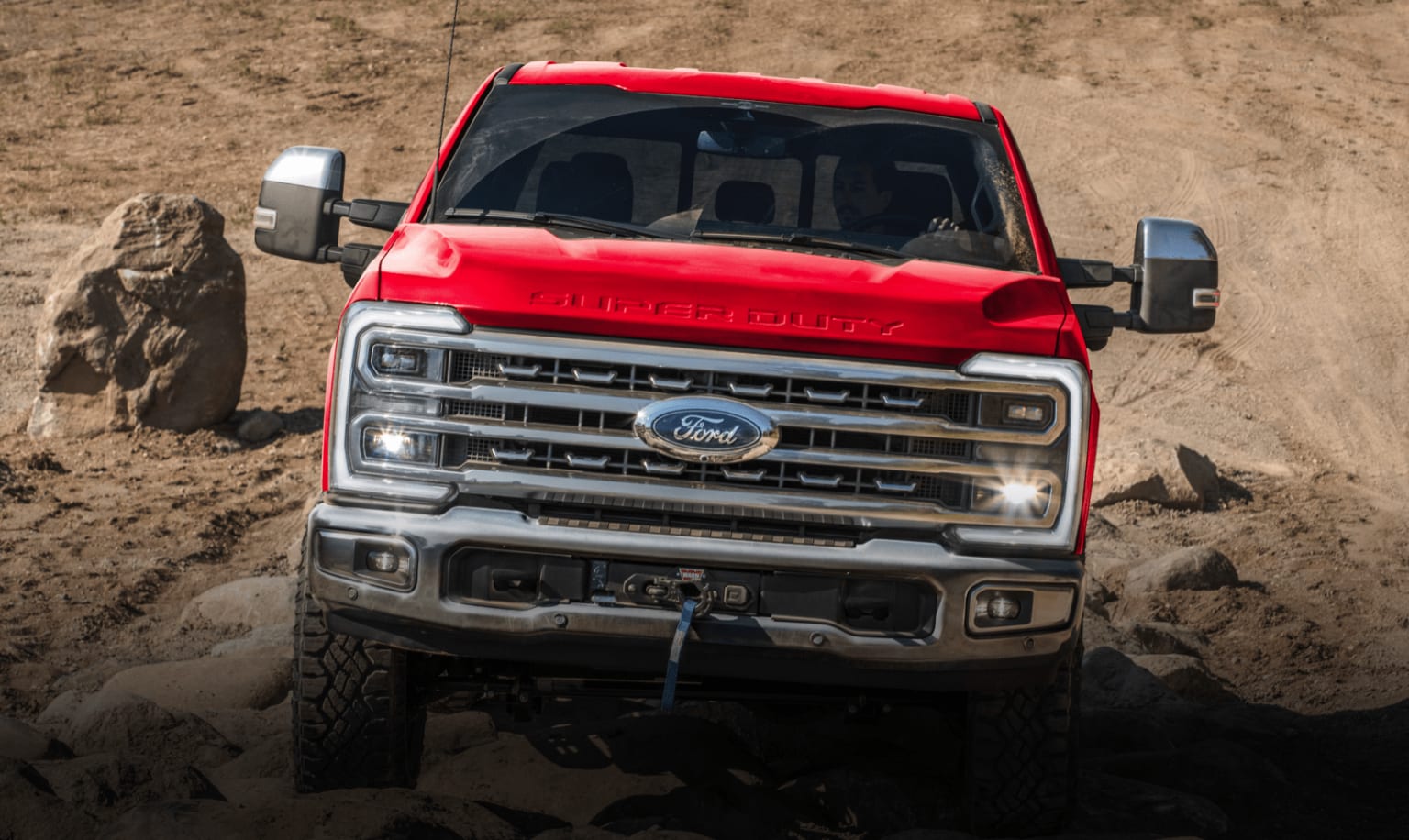 A red truck is navigating through a rugged, sandy terrain with large rocks in the background.