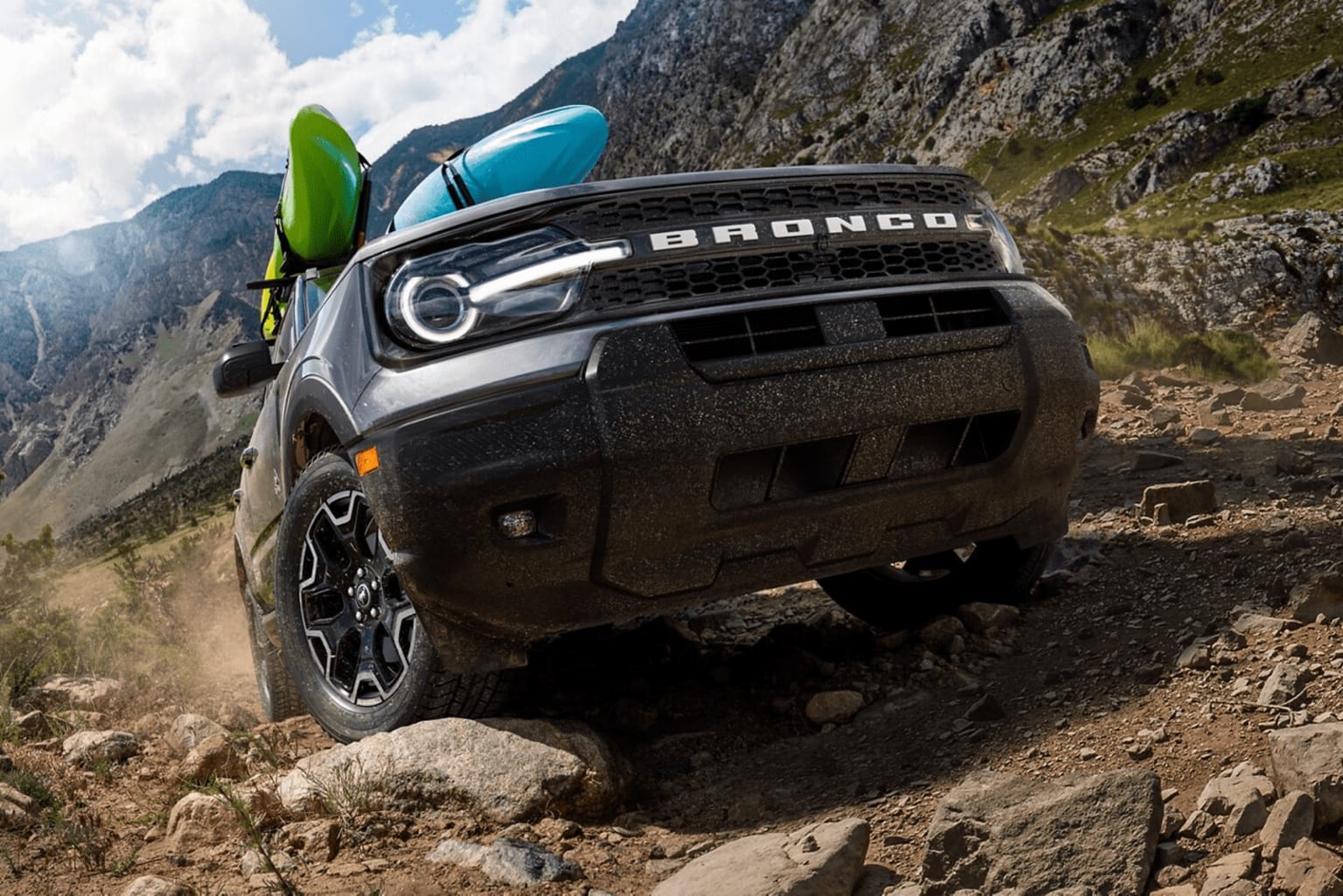A rugged off-road vehicle navigates a rocky, mountainous terrain with a backdrop of towering cliffs and a cloudy sky.