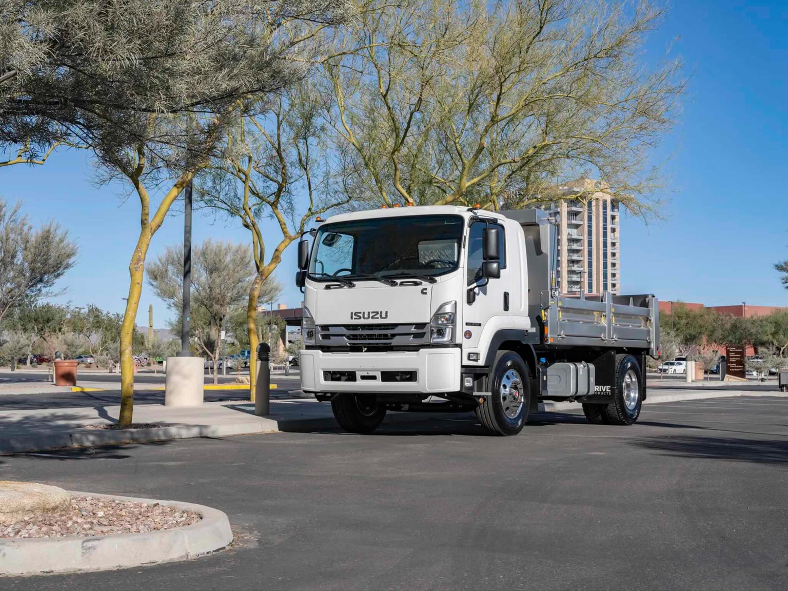 A white flatbed truck is parked on an asphalt lot in the foreground, with sparse trees and a building visible in the background under a clear blue sky.