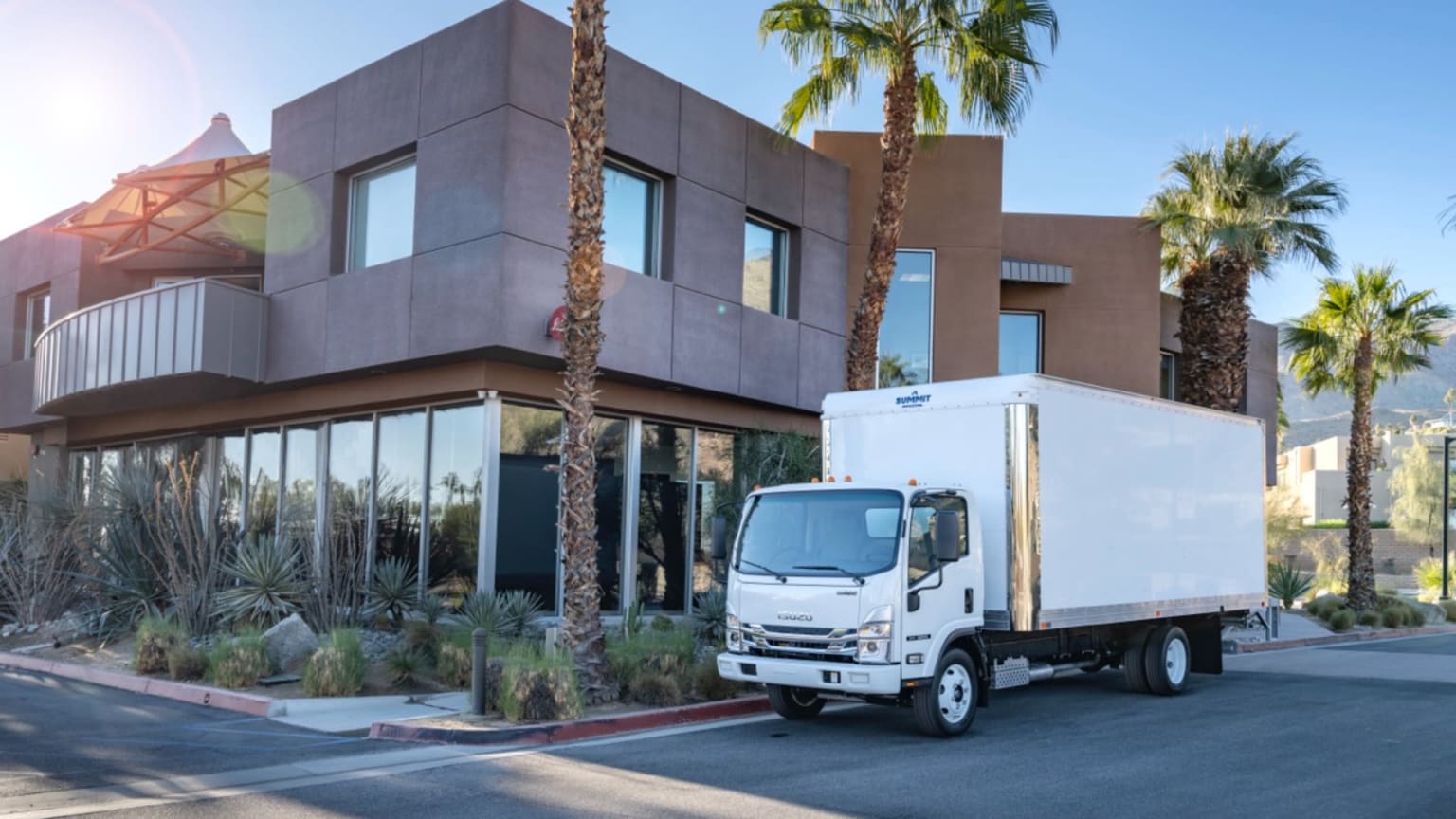 A modern automotive dealership building with large windows and palm trees in the foreground, with a white delivery truck parked in the driveway.