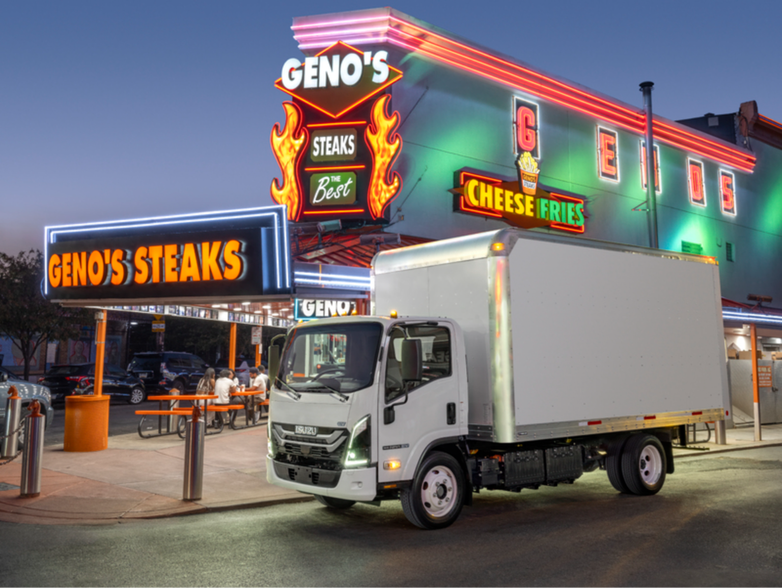 A white box truck parked in front of a brightly lit restaurant at dusk.