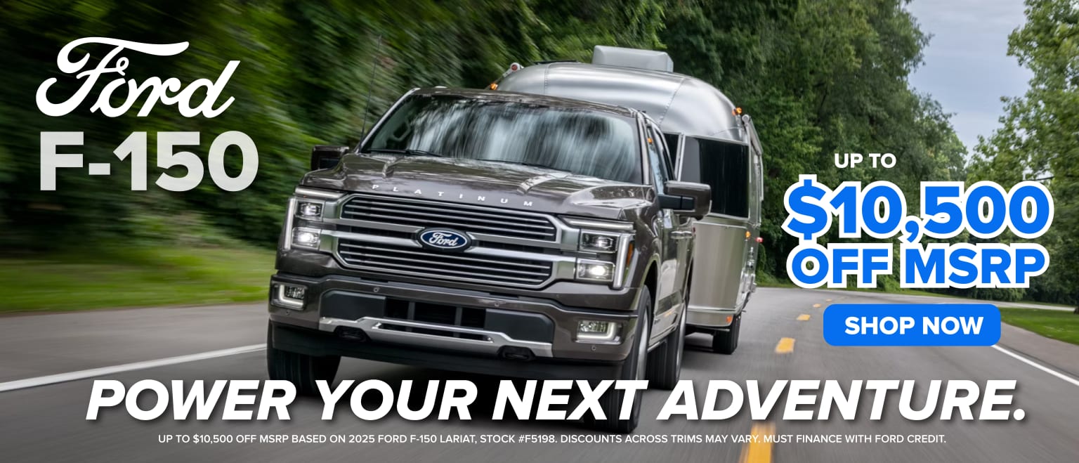 The image shows a large, silver pickup truck driving on a road surrounded by lush, green trees. The truck is prominently featured in the foreground.