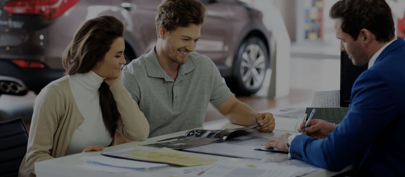 A couple seated at a desk speaking to a salesperson in an automotive dealership showroom.