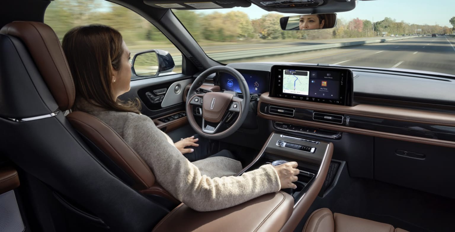 Interior view of a vehicle with a person in the driver's seat and a view of the road through the windshield.