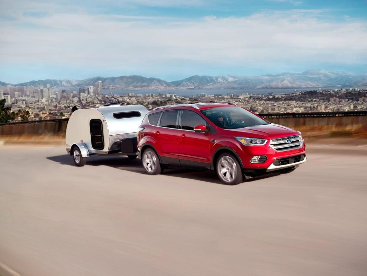 A red SUV towing a small camper trailer driving on a road with a cityscape and mountains in the background.