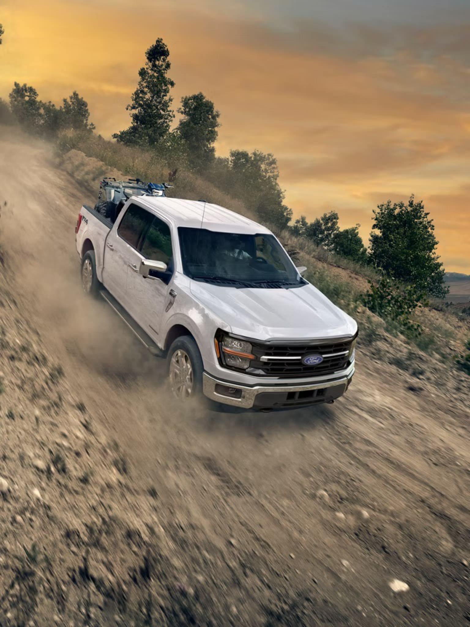 A white pickup truck is speeding through a dusty, rugged terrain against a backdrop of a vibrant sunset sky and lush, forested landscape.
