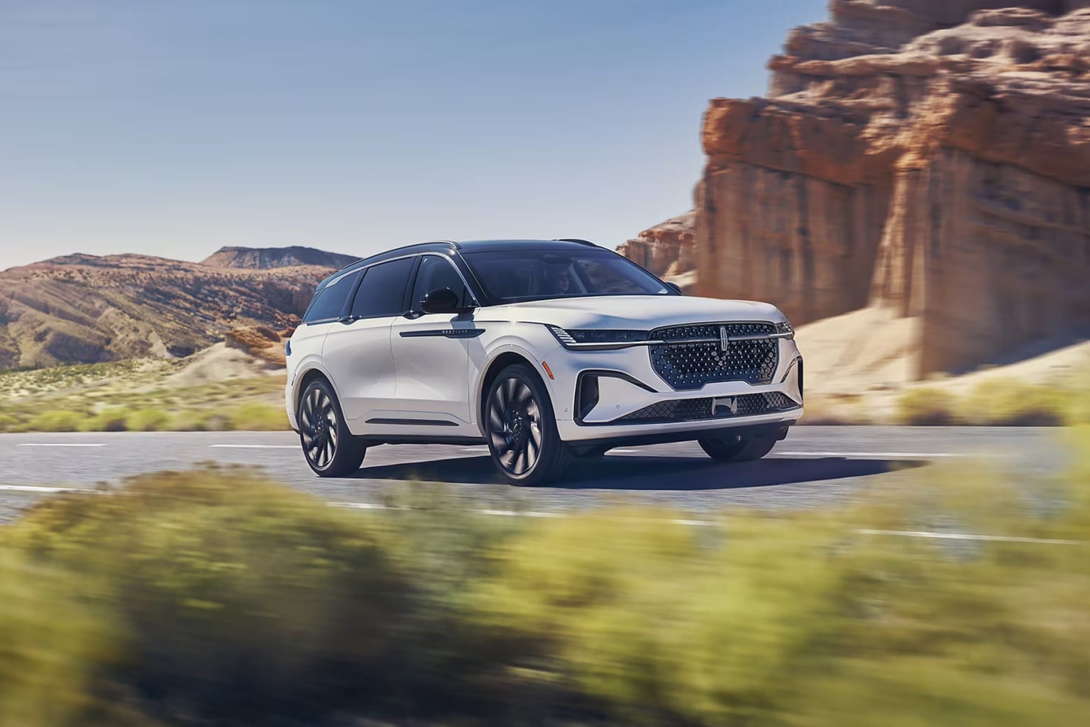 A white SUV driving on a winding road through a rugged mountainous landscape with rocky cliffs and lush vegetation in the background.