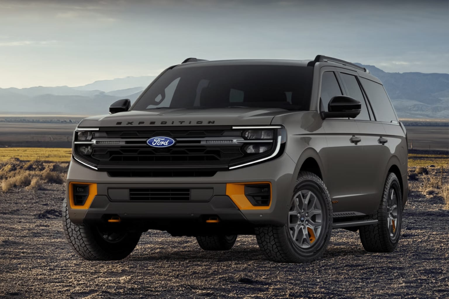 A gray SUV with orange accents parked on a dirt road in front of a vast, arid landscape with mountains in the distance.