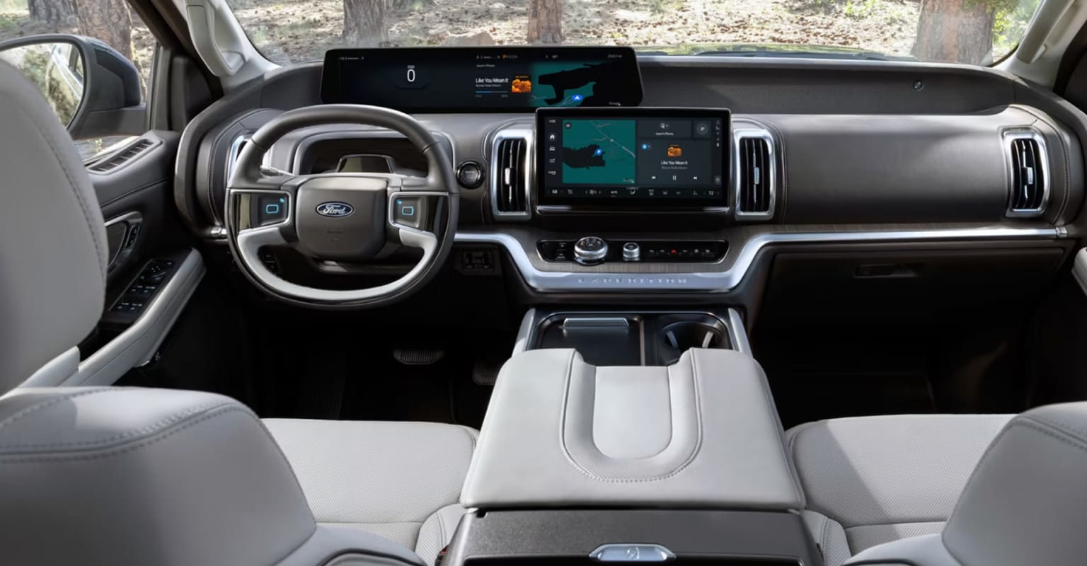 The image shows the interior of a modern vehicle with a large touchscreen display and various controls and features visible in the foreground. The background appears to be a wooded area, suggesting an outdoor setting.