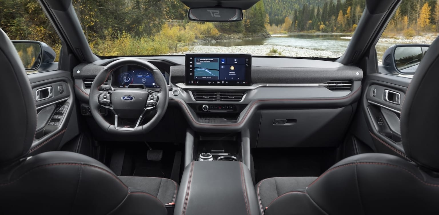 Interior of a modern vehicle with a large touchscreen display, steering wheel, and dashboard controls, showcasing a scenic outdoor landscape with a river and trees visible through the windshield in the background.