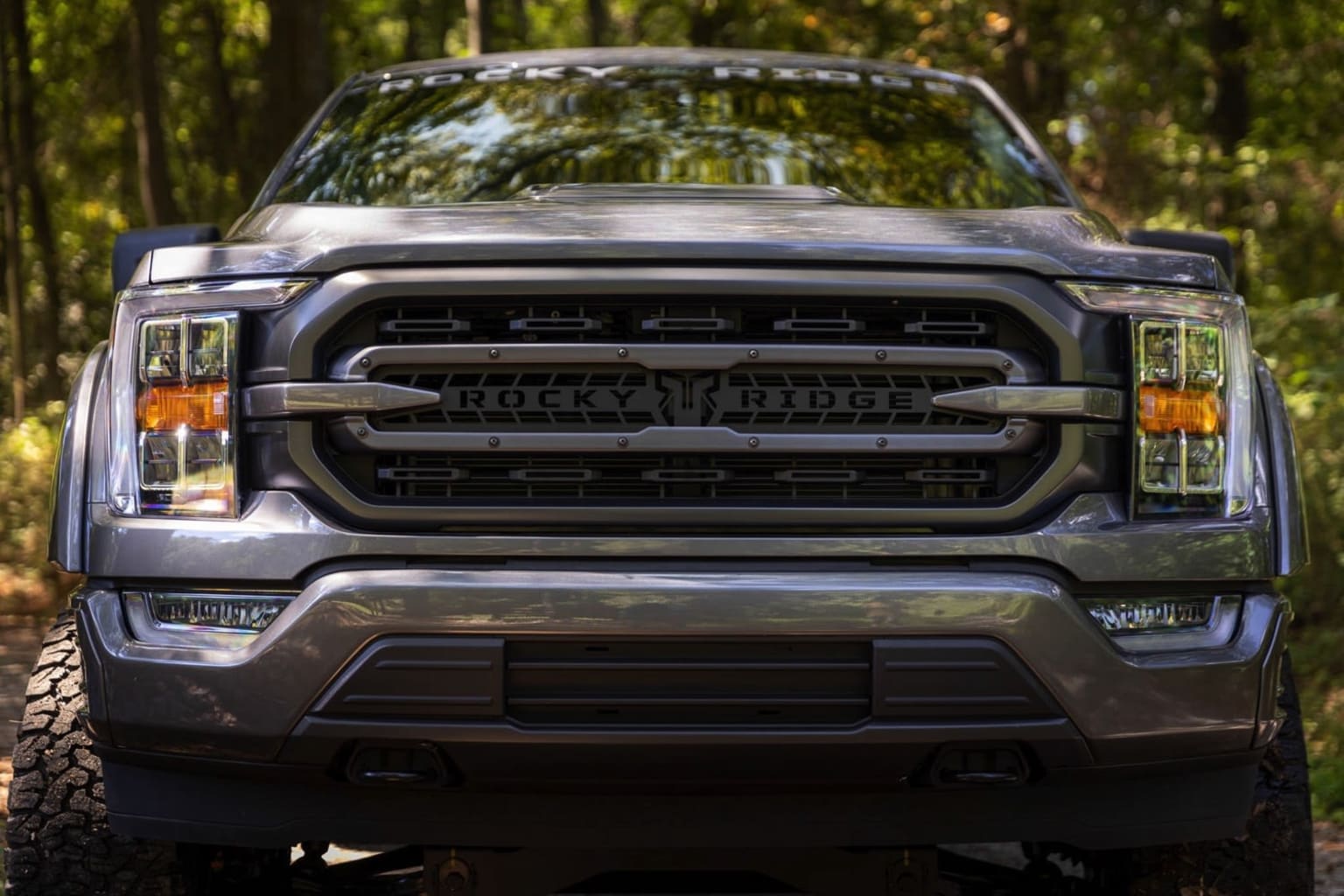 A rugged and powerful-looking truck with custom branding prominently displayed on the front grille, set against a backdrop of lush, green foliage.