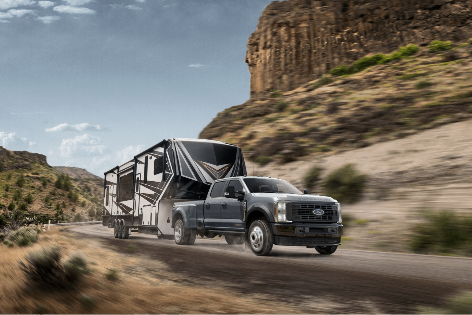 A dark gray truck tows a large, modern RV down a dusty road with rocky, arid hills in the background.