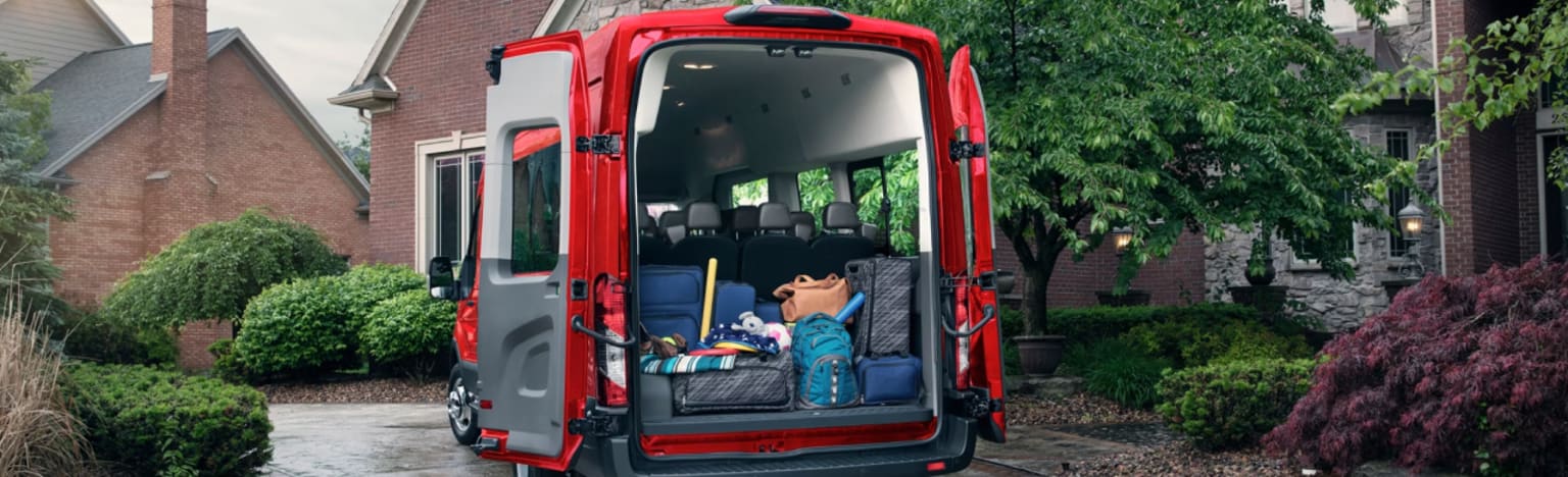 A red van filled with luggage parked in a driveway with houses and trees in the background.