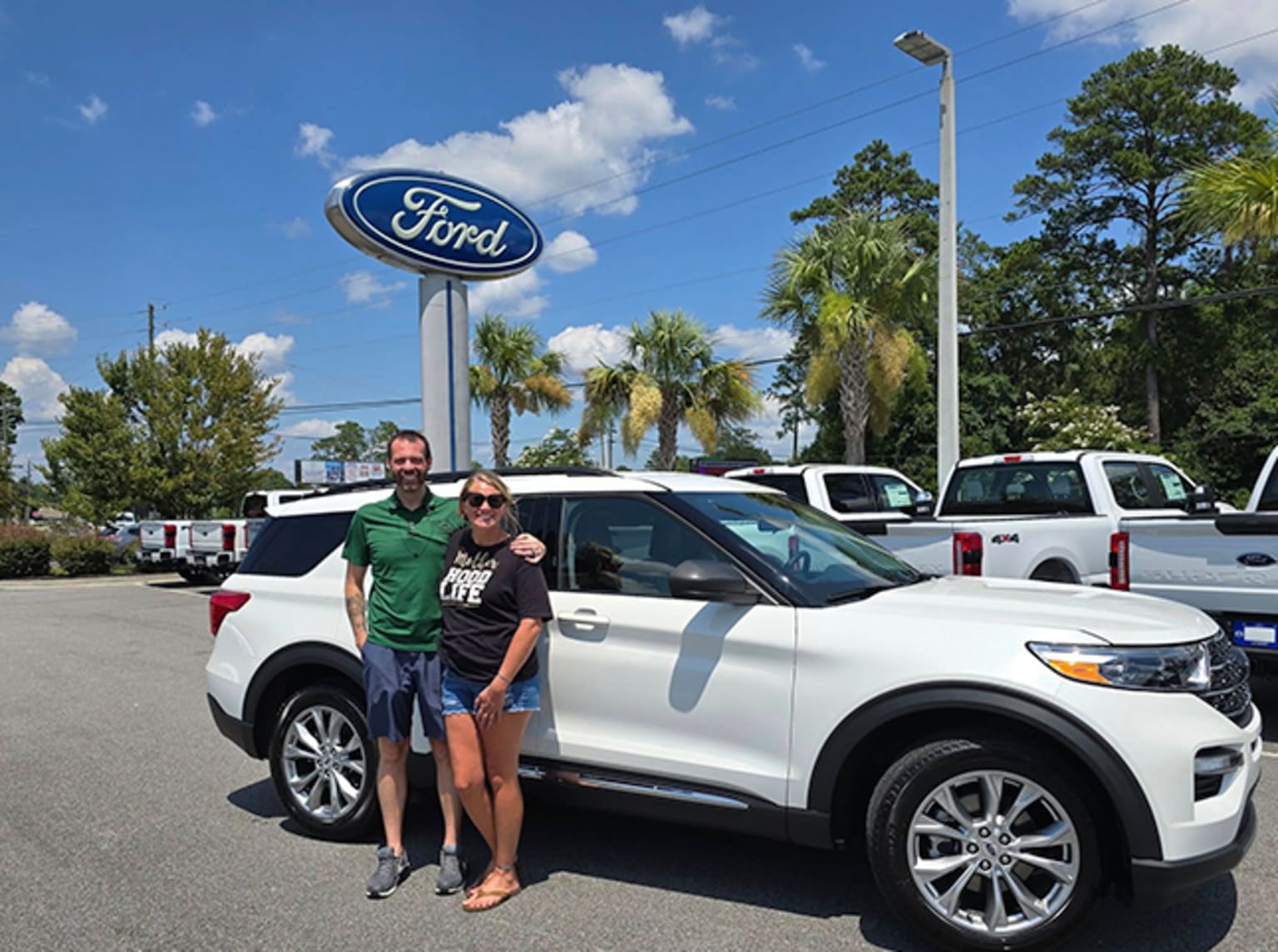 The image shows a couple standing in front of a white SUV in a dealership parking lot, with palm trees and a blue sky in the background.