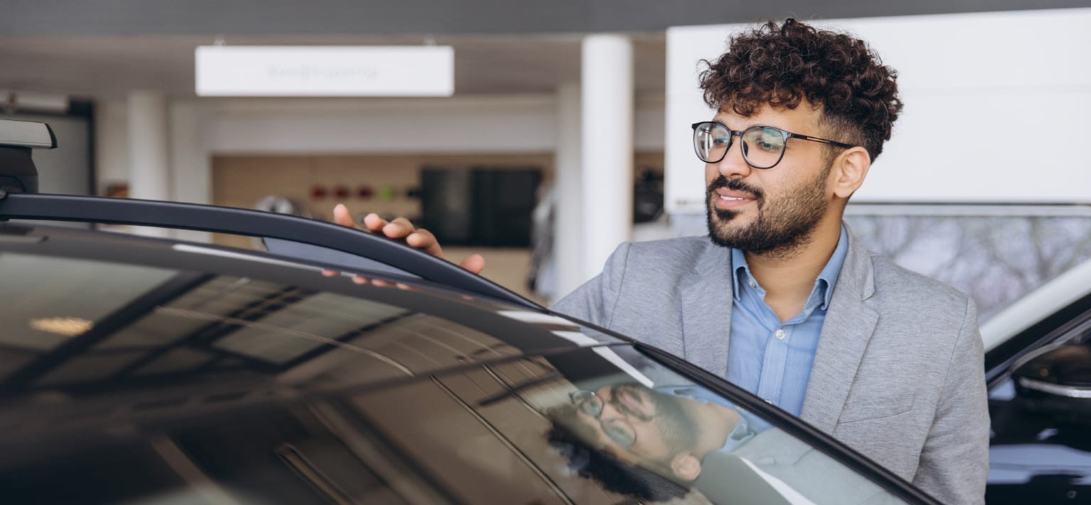 A man in a suit and glasses inspecting the roof rack of a car in a showroom.