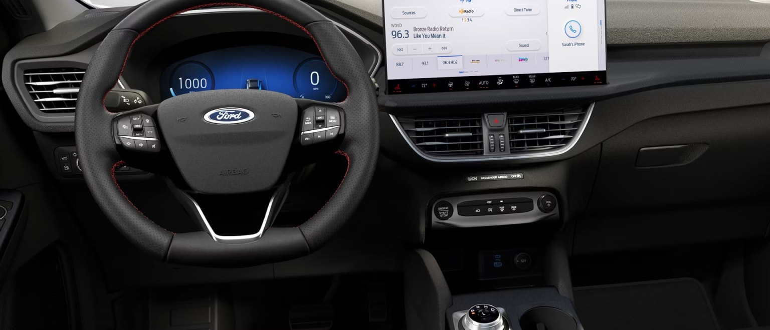 The image shows the interior of a modern vehicle, with a steering wheel, dashboard, and a large touchscreen display in the center console. The dashboard displays various information and controls, indicating this is a technologically advanced car.