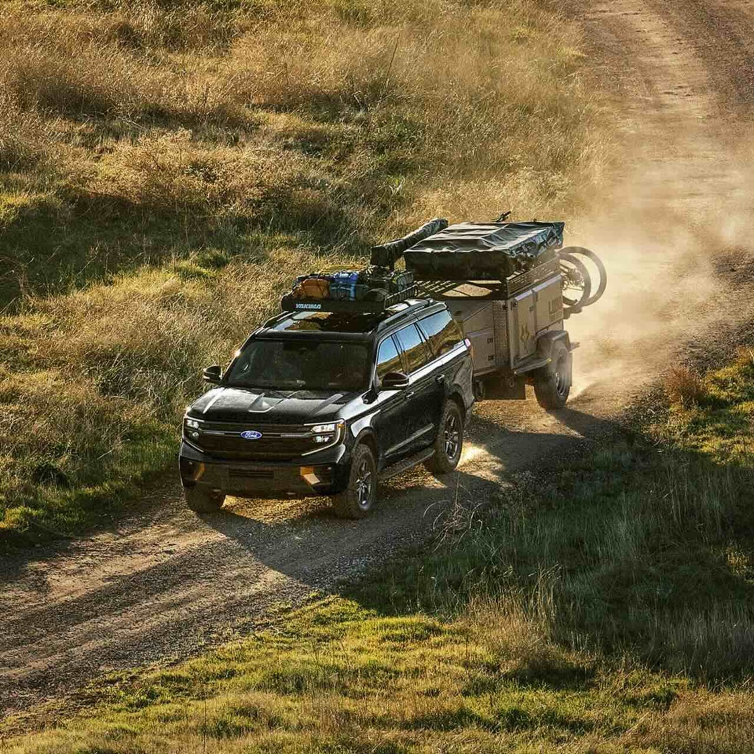 Black SUV towing a trailer on a dusty dirt road with dry, grassy hills in the background.