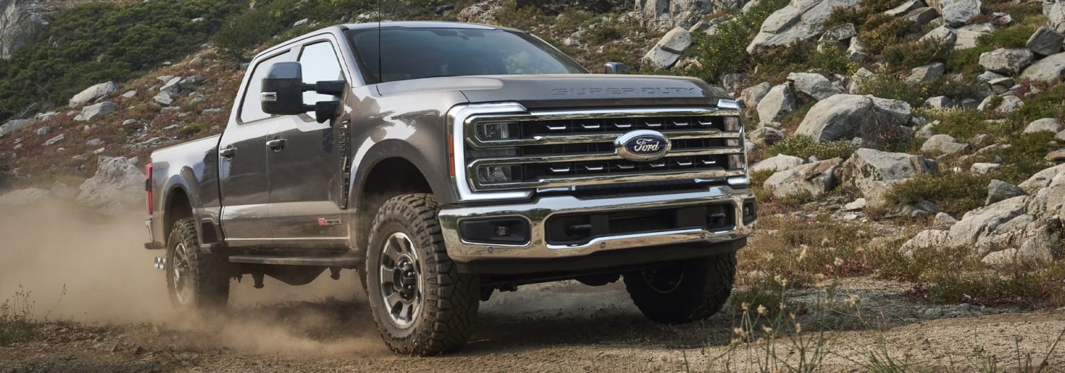 A large pickup truck is driving through rocky, mountainous terrain, kicking up a cloud of dust behind it.