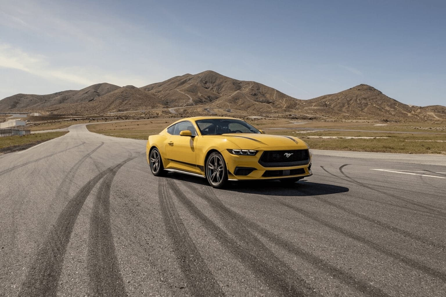 The image shows a yellow sports car driving on a road in a desert landscape, with rugged mountains in the background.