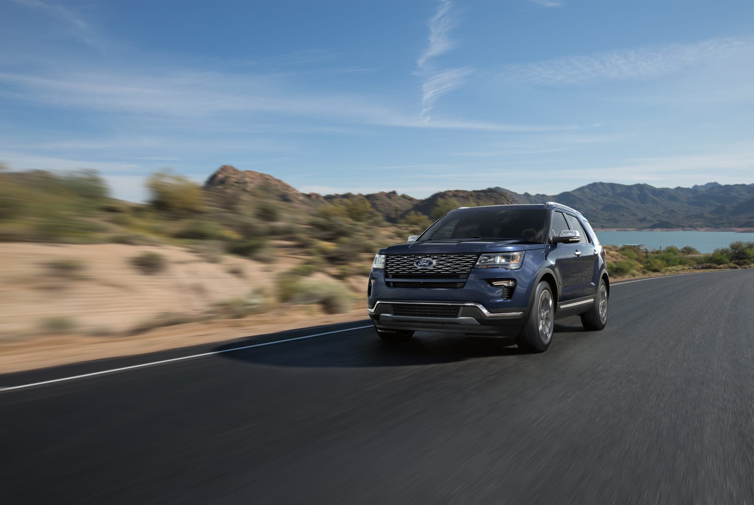 a blue suv driving down a road next to a body of water with mountains in the background and a blue sky