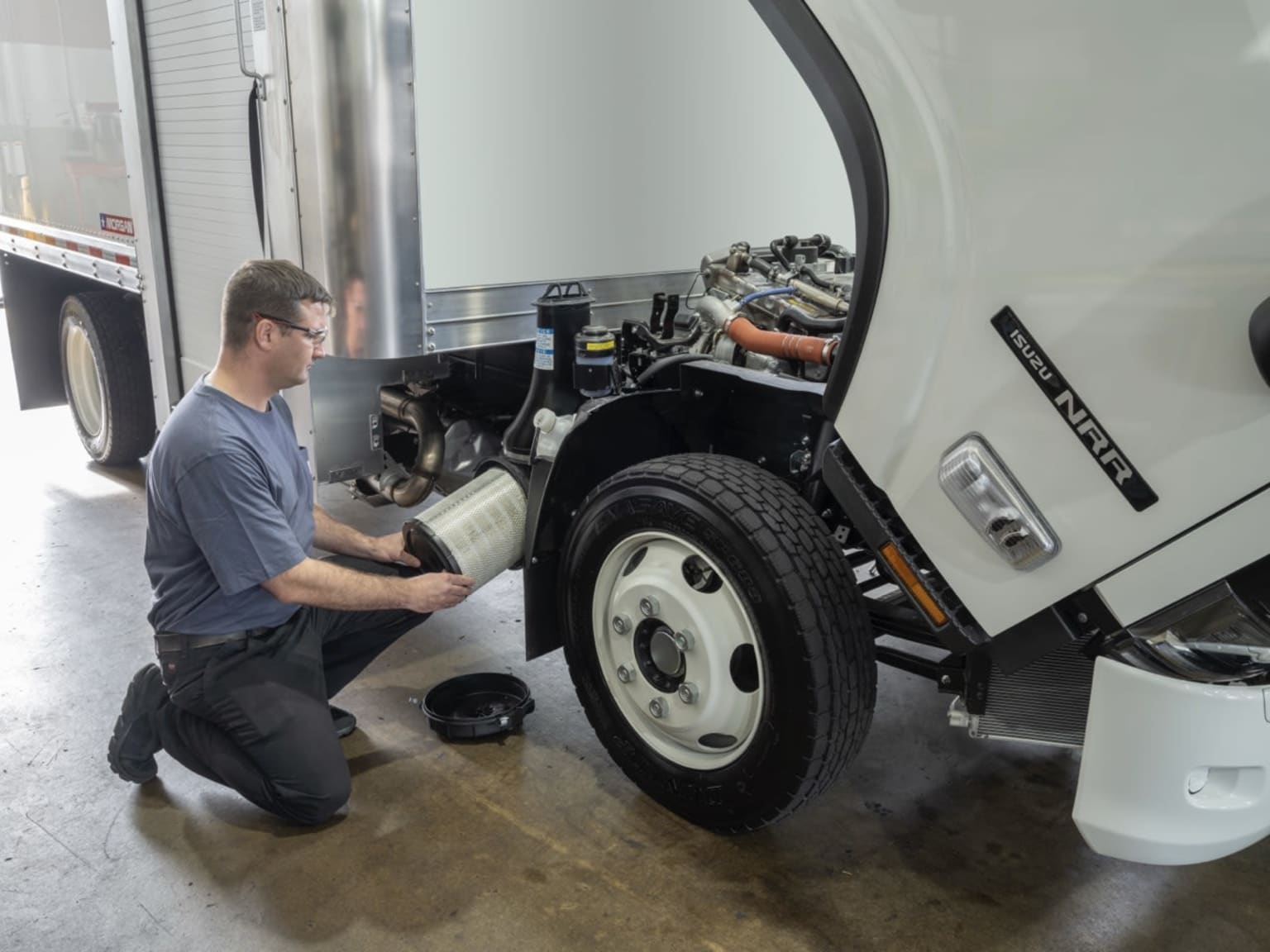 A person in a blue shirt inspecting a large truck's tires and components in a garage or workshop setting.