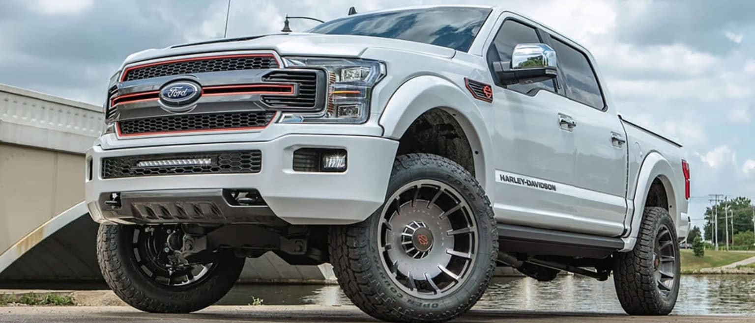 A white truck with custom wheels and off-road tires is parked near a body of water with a bridge in the background.