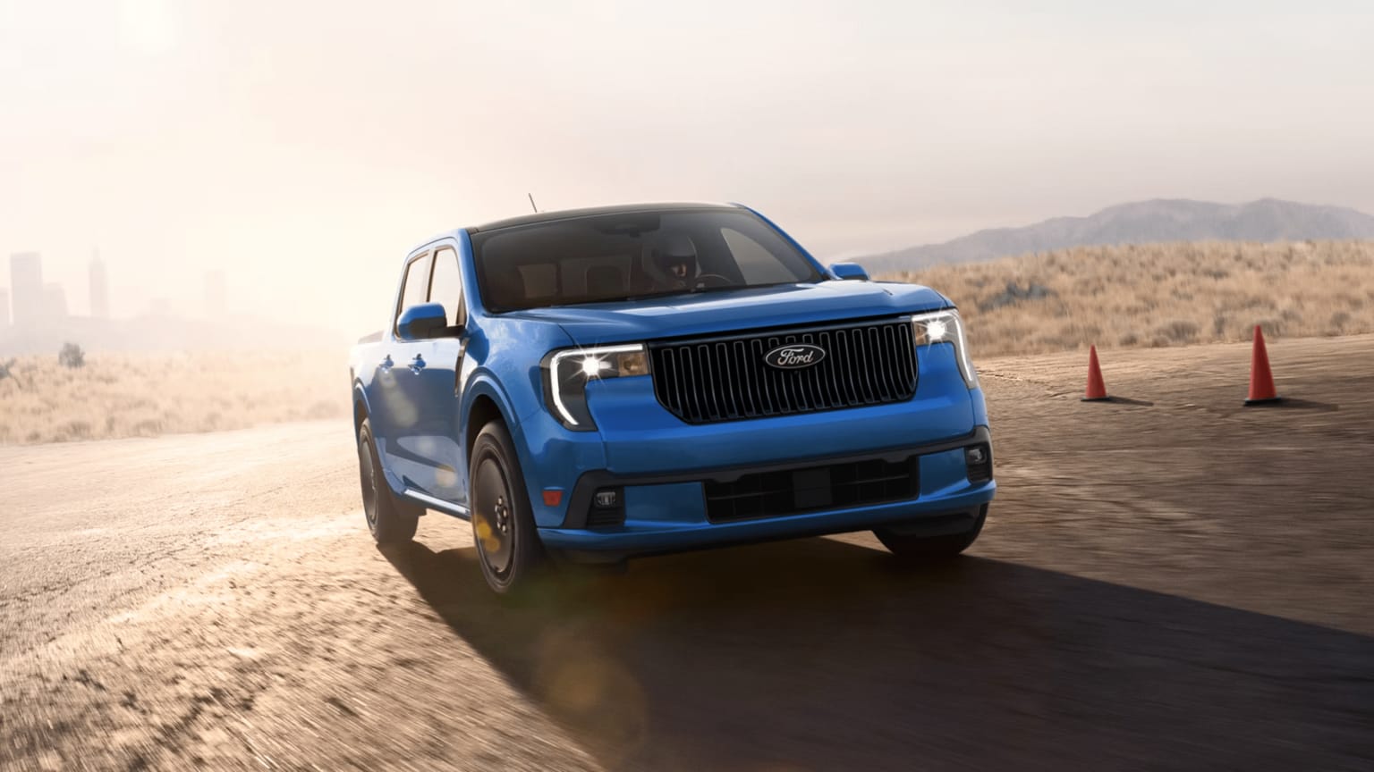 Blue electric truck driving in desert landscape with traffic cones and mountains.