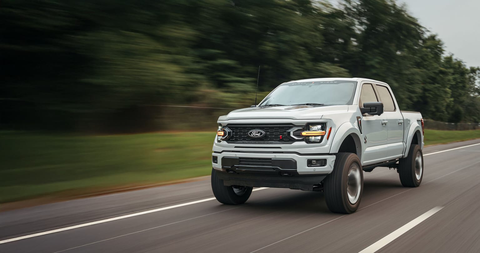 A white pickup truck with large off-road tires speeding down a road surrounded by blurred trees and foliage, creating a sense of motion and speed.