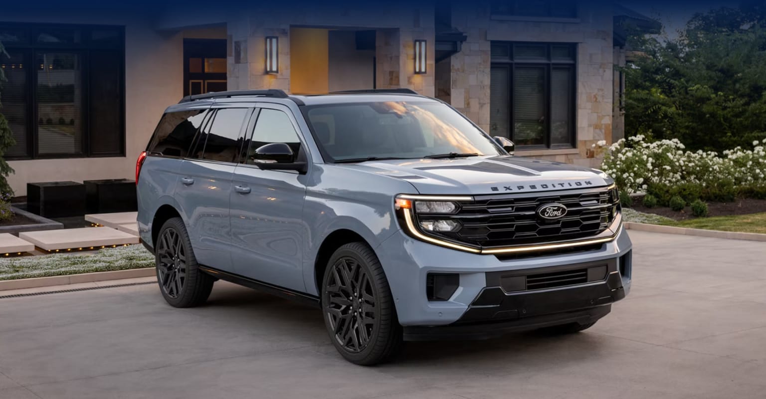 A large, silver SUV with a prominent grille and sleek design is parked in front of a modern, multi-story building surrounded by lush greenery.