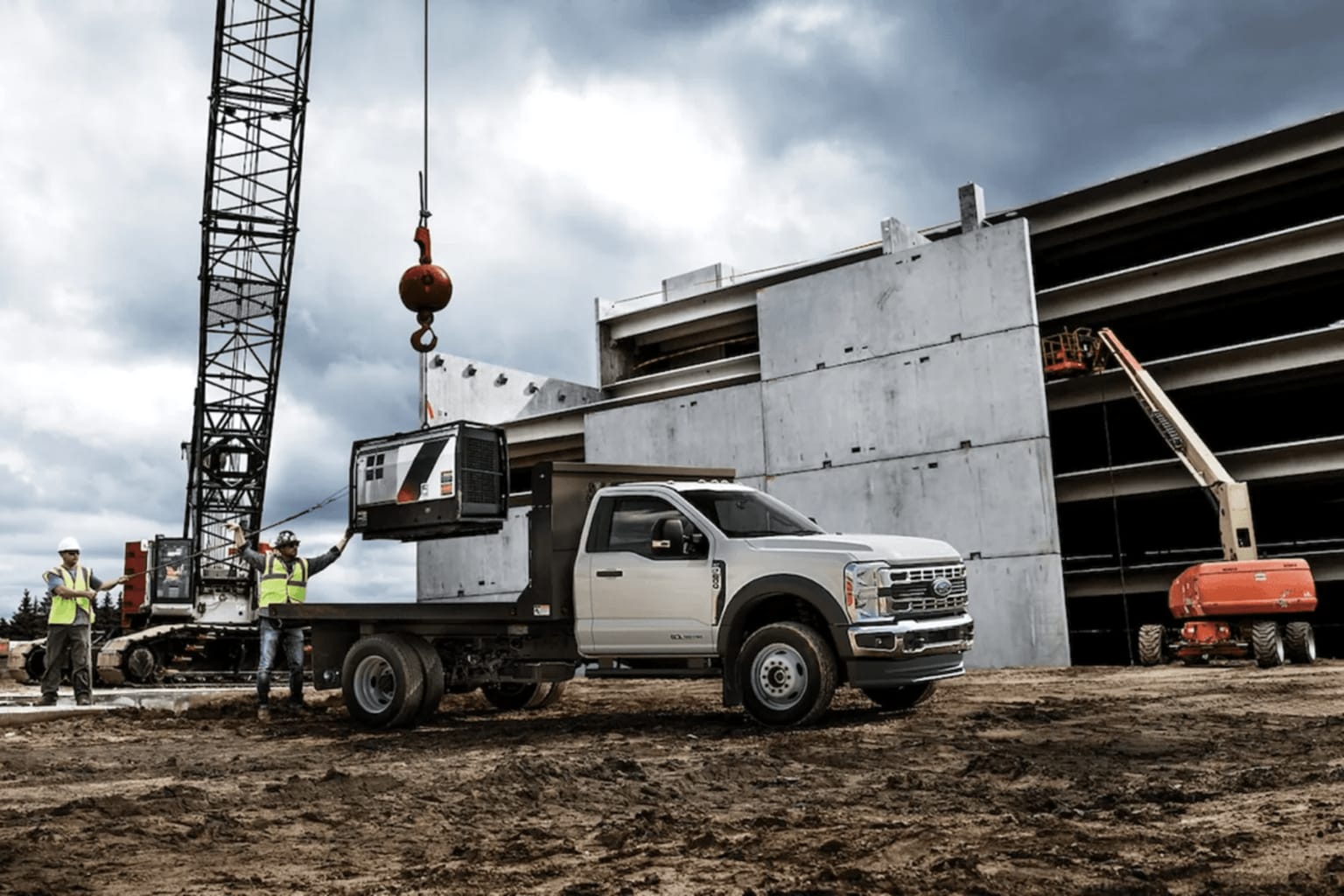 A large construction crane towers over a work site, where a heavy-duty truck is parked on the muddy ground, surrounded by construction materials and workers in safety gear.