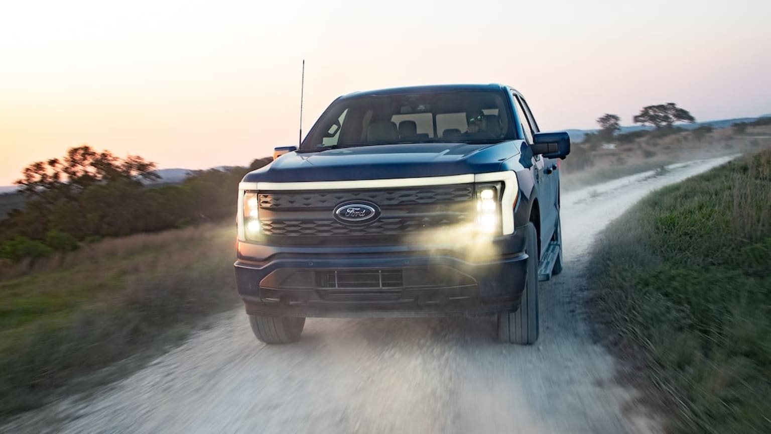 a truck driving down a dirt road near a grassy field with trees and bushes in the background at sunset