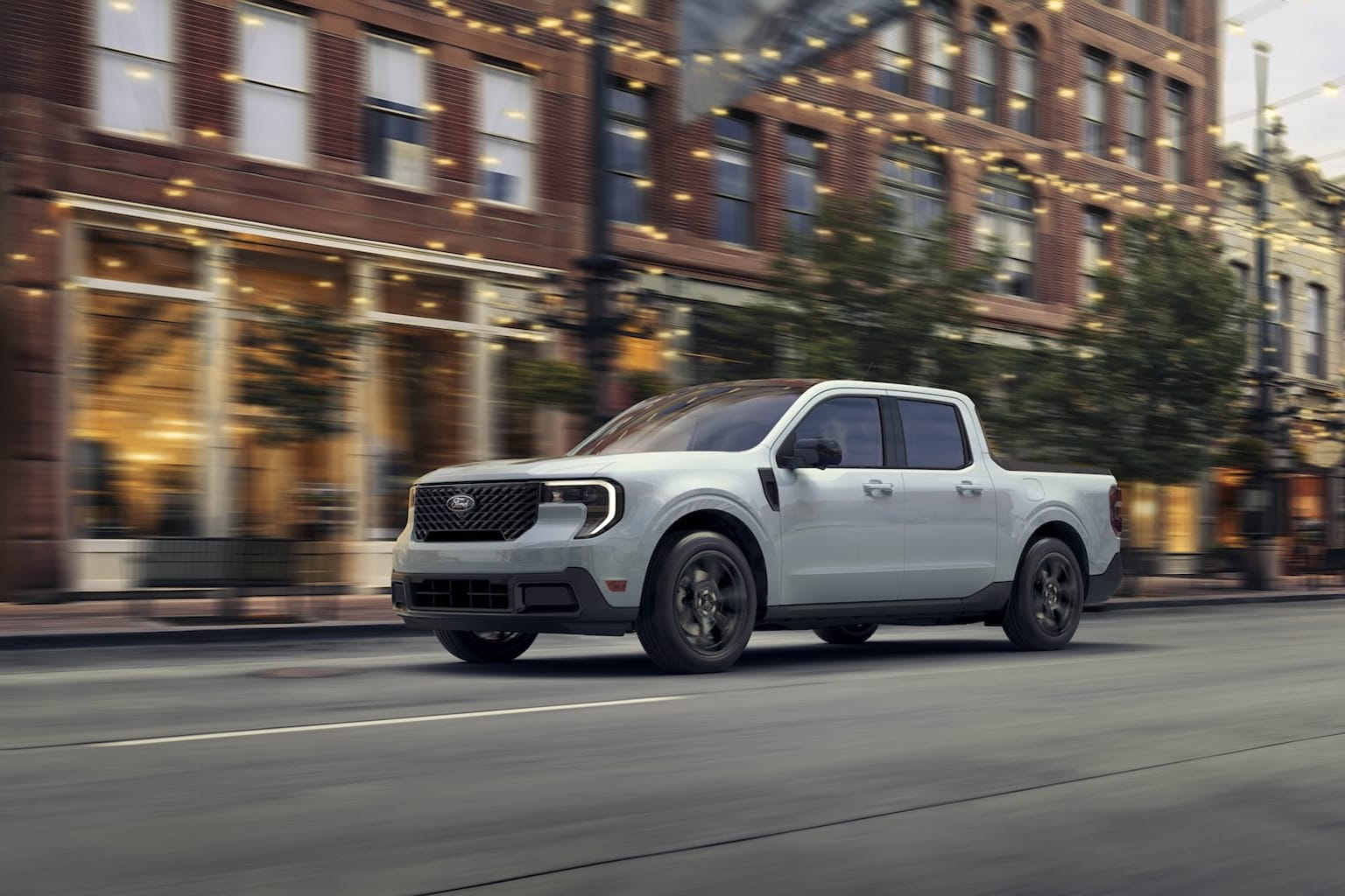 A white pickup truck driving down a city street lined with brick buildings and festive string lights, creating a vibrant and lively urban scene.