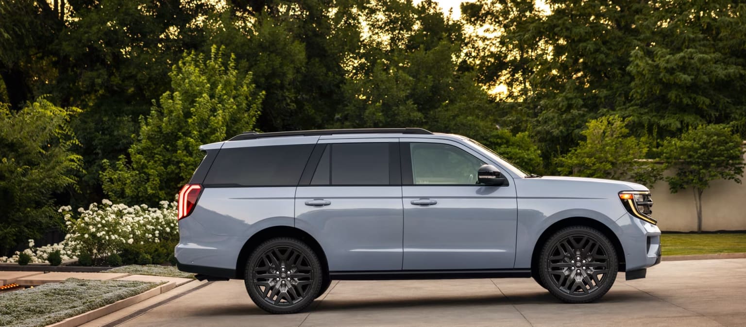 White SUV parked on a driveway with greenery and trees in the background.