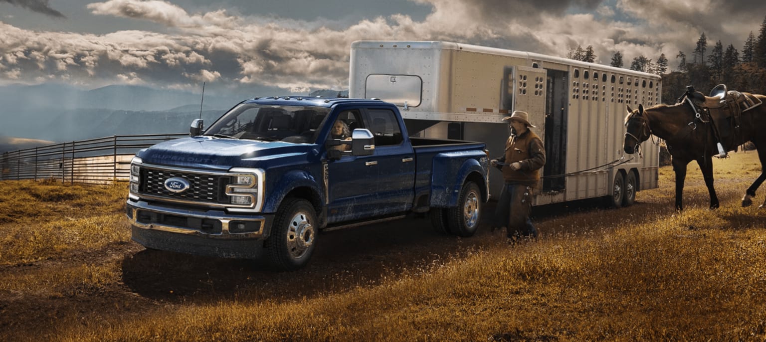 A rugged blue pickup truck towing a large trailer is parked in a grassy field, with a horse standing nearby and dramatic cloudy skies in the background.