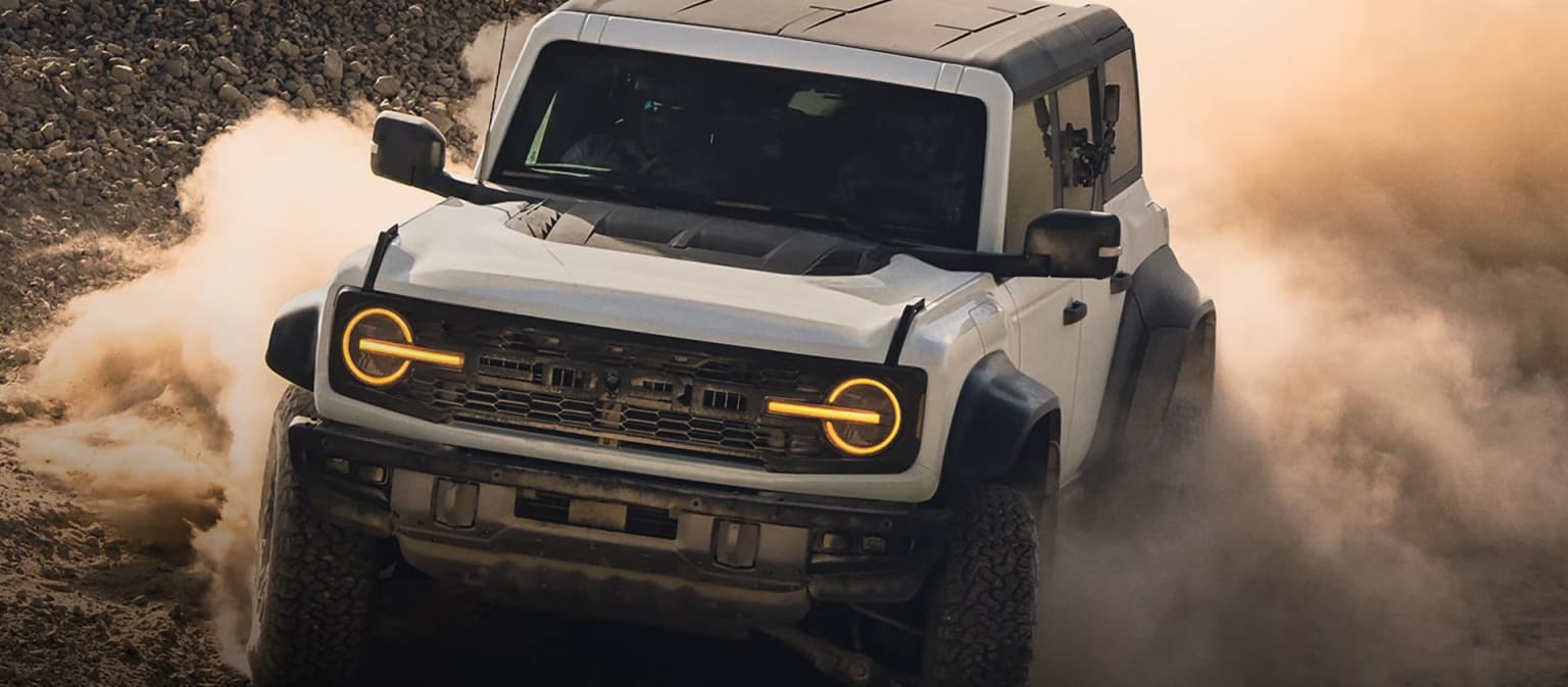 A white SUV kicks up dust as it drives on a dirt road with a hazy, orange sky in the background.