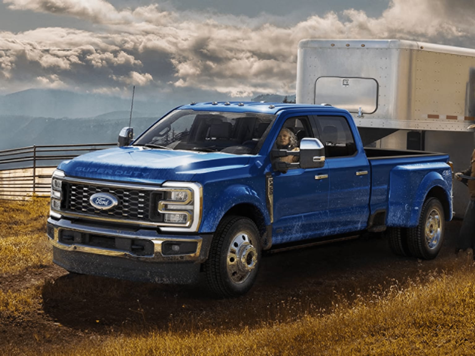 Blue truck parked on grass field with silver trailer and mountains in background