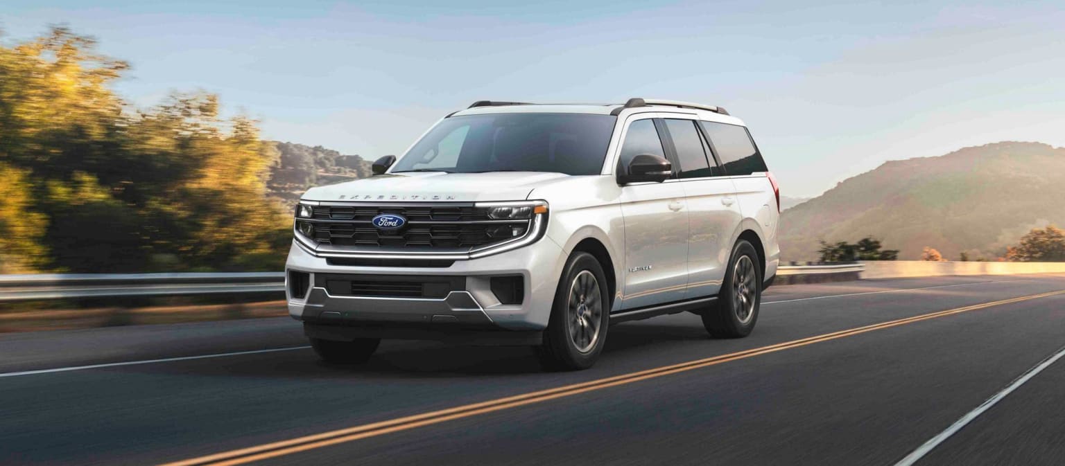 A large, white SUV driving on a winding mountain road surrounded by lush, forested hills in the background.