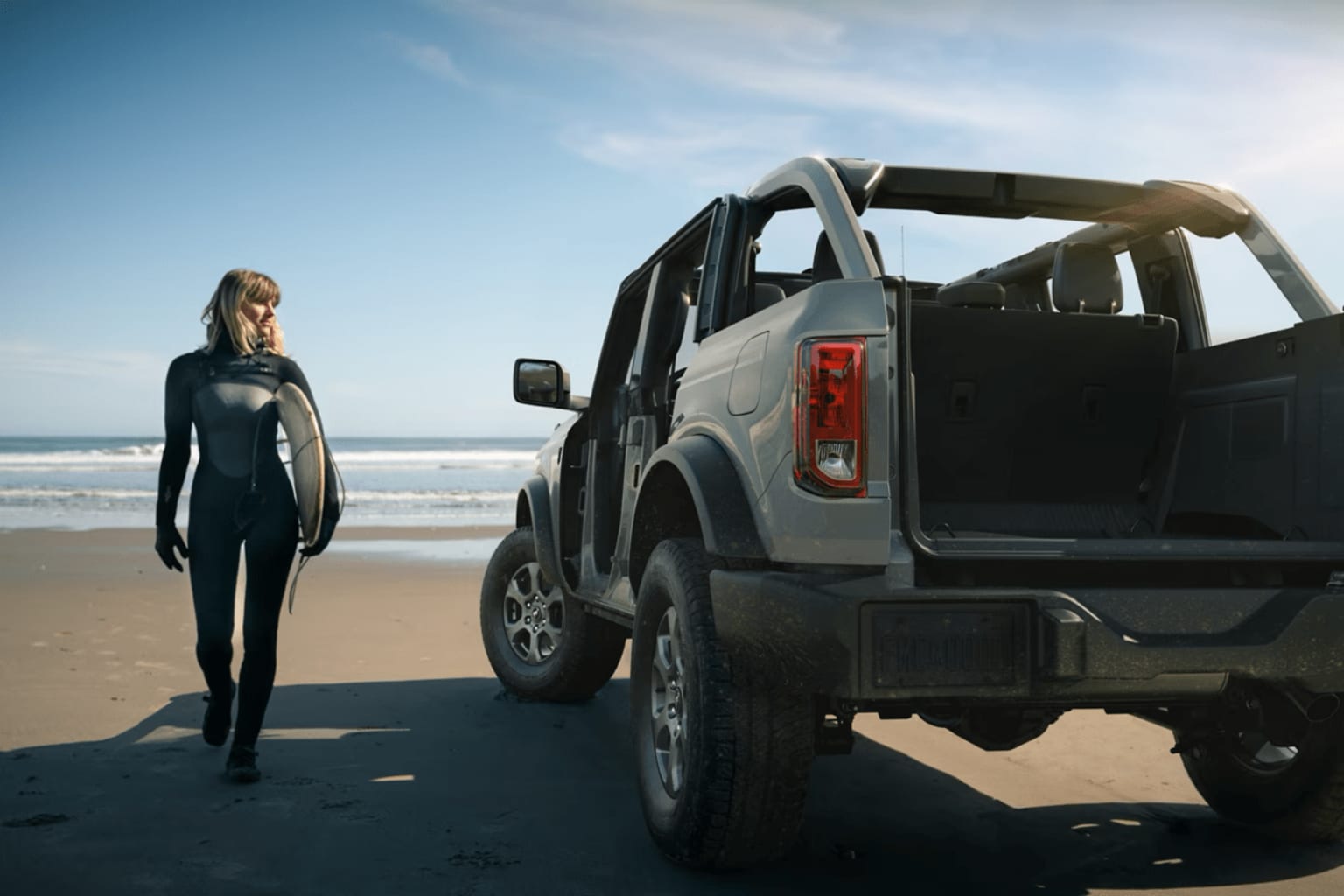 A person in a black wetsuit stands next to a large off-road vehicle on a sandy beach, with the ocean visible in the background.
