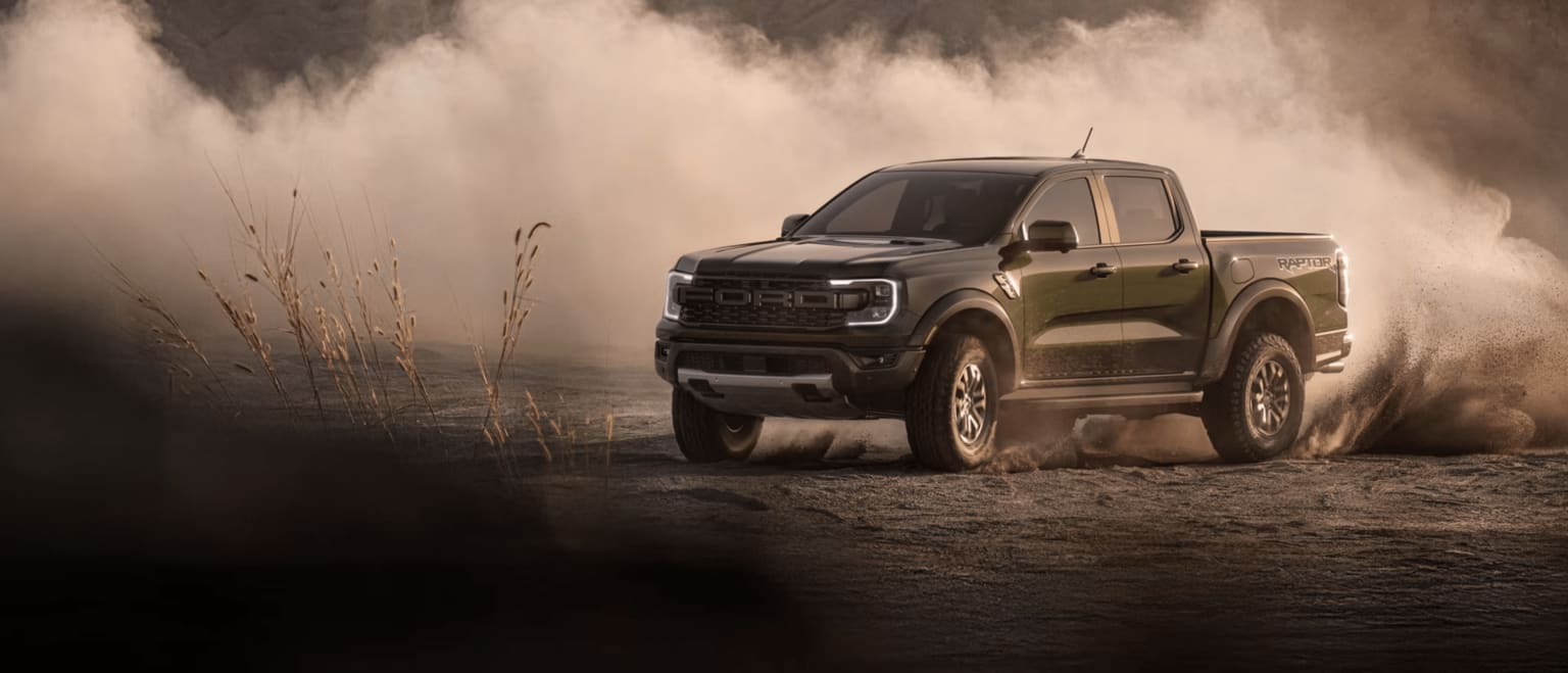 A dark green truck kicks up a cloud of dust as it drives through a dry, barren landscape.