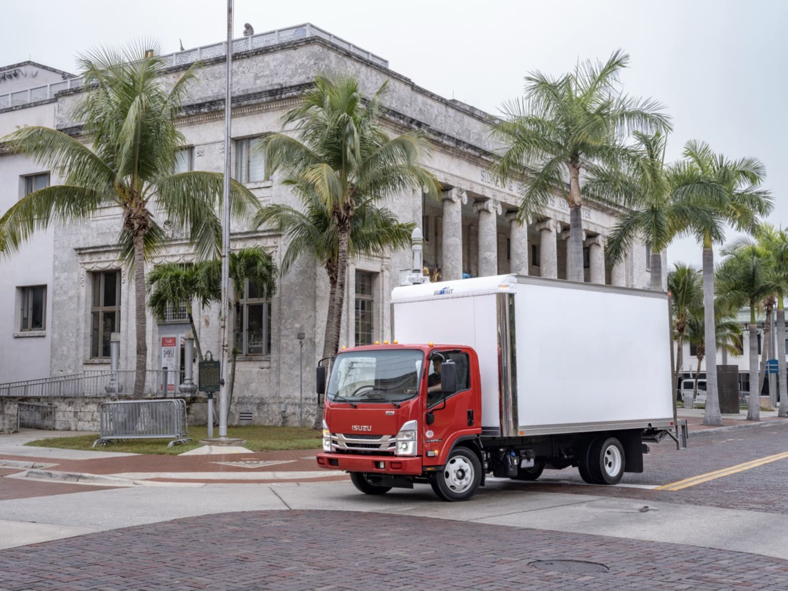 A red box truck drives past a grand building with columns and palm trees.