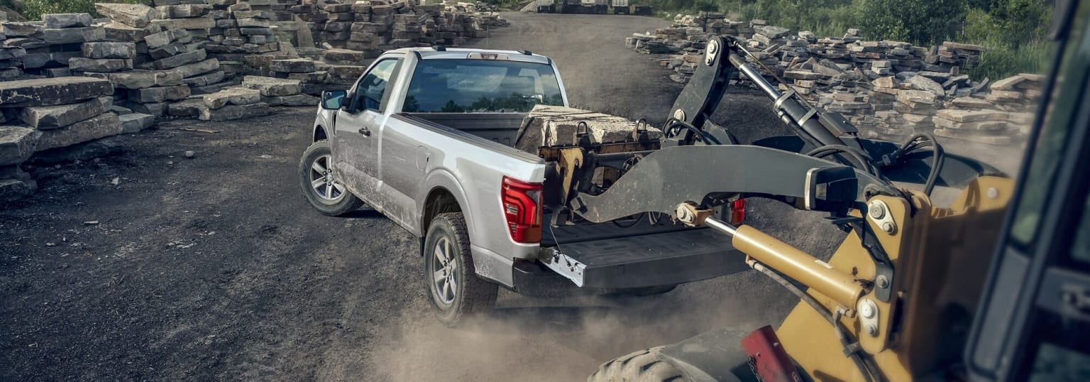 A white pickup truck with a flatbed in the foreground, surrounded by a cluttered background of stacked construction materials and equipment.