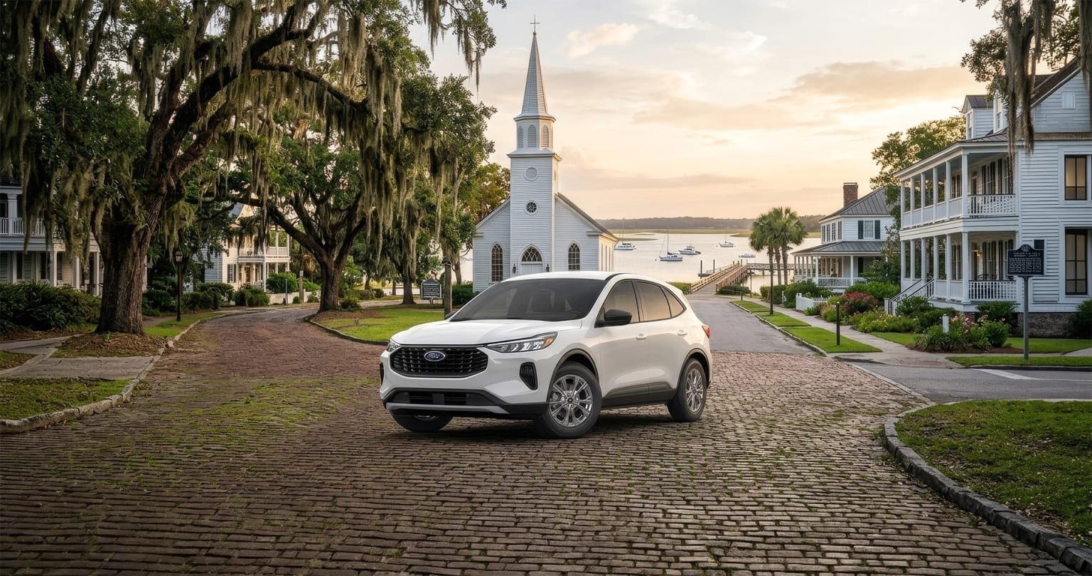 White SUV parked on cobblestone street in front of historic buildings and church steeple, surrounded by lush greenery.