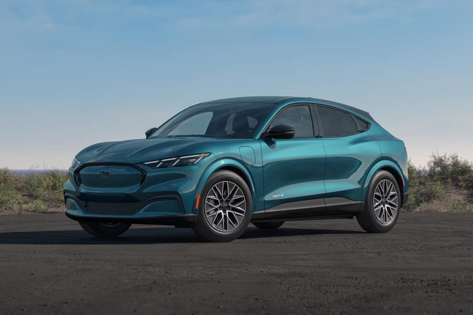 A teal electric SUV is parked on a dirt road with a clear blue sky and distant hills in the background.